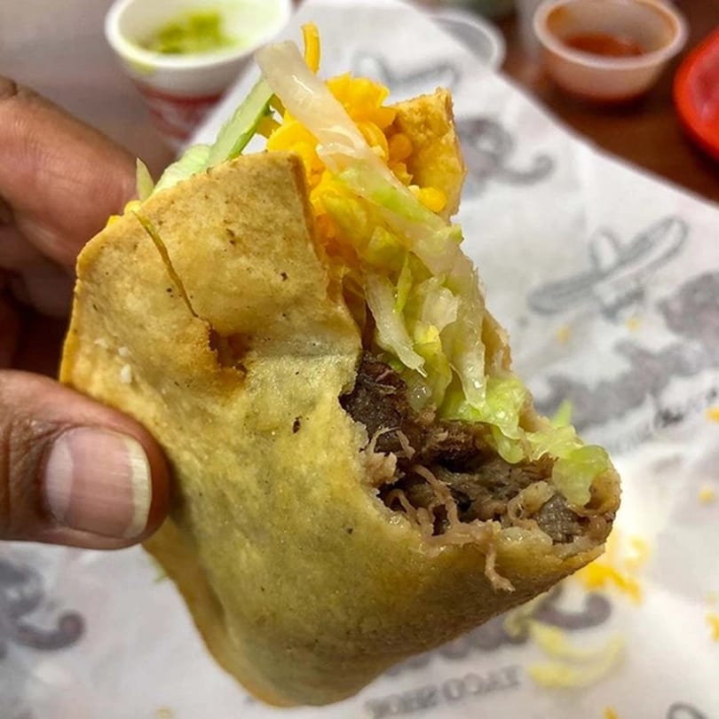 A hand holds a partially eaten Roberto's Taco Shop taco filled with meat, cheese, and lettuce.
