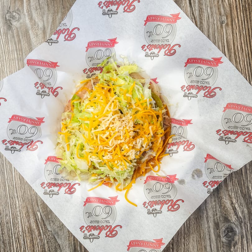 A Roberto's Taco Shop tostada with shredded lettuce and cheese on branded paper.